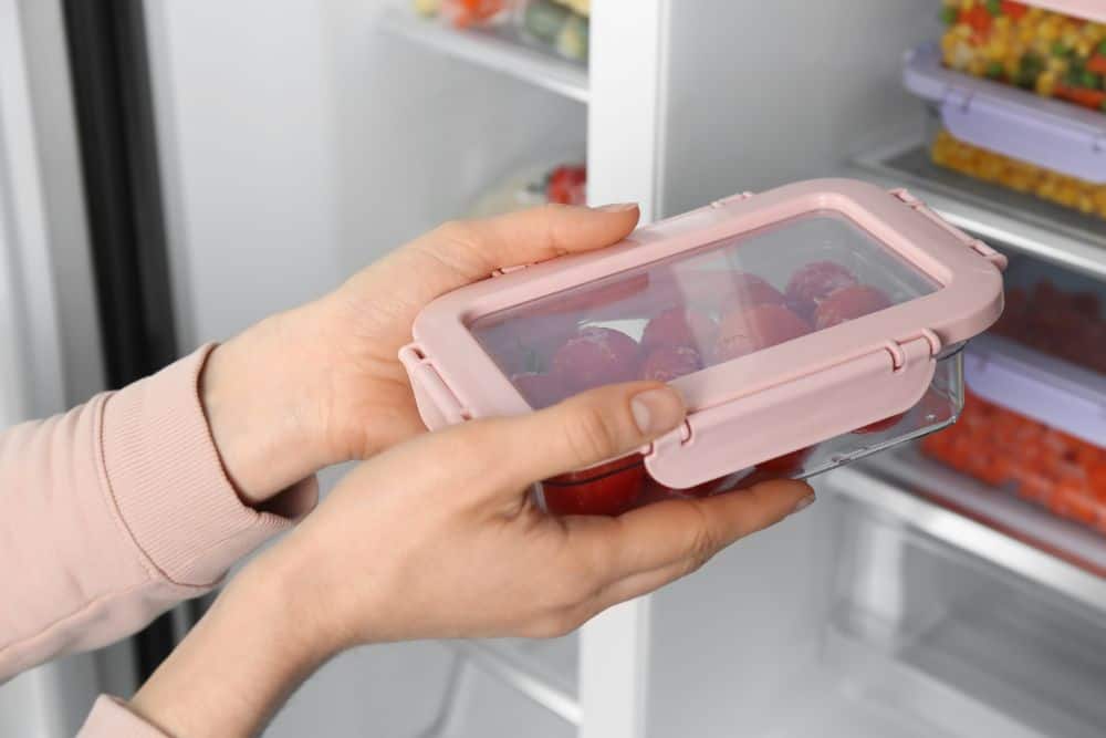 A person checking if glass can explode in the freezer, holding a plastic container in an open refrigerator.